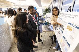 A student points at her research poster, explaining her work to two listeners